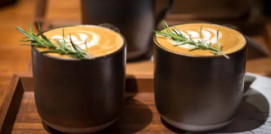 Dos tazas de café con arte latte y una rama de romero como decoración, servidas sobre una bandeja de madera en un ambiente de cafetería.