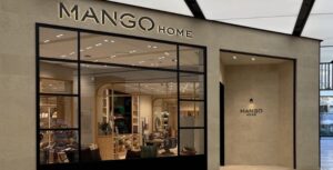 Primer plano de la fachada de una tienda Mango Home en un centro comercial, con paredes de piedra caliza lisa y grandes ventanales con marcos negros. El interior está iluminado y se ven muebles y decoración.