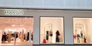 Vista de la entrada y tres grandes escaparates de una tienda de ropa Mango. El interior está pintado de rosa pastel y bien iluminado. Maniquíes con la colección actual en los escaparates.